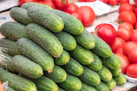 A stack of fresh cucumbers displayed at an outdoor farmers market with ripe tomatoes.